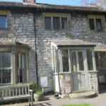 Old stone cottage with a small porch, large bay window, wooden bench, and a garden filled with red flowers in front. Sunlight highlights the textured stone walls and greenery, creating a welcoming retreat—perfect as a Christian holiday cottage nestled in the picturesque Peak District.
