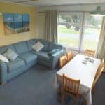 A cosy living-dining room in this Christian holiday chalet features a blue sofa with patterned cushions, a wooden dining table with six chairs, large windows with curtains, and a beach painting on a yellow wall—perfect for relaxing after exploring beautiful Pembrokeshire.