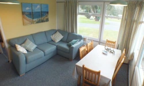 A cosy living-dining room in this Christian holiday chalet features a blue sofa with patterned cushions, a wooden dining table with six chairs, large windows with curtains, and a beach painting on a yellow wall—perfect for relaxing after exploring beautiful Pembrokeshire.