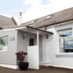 Small white and grey house with a sloping roof, front porch, and a potted plant near the entrance, set against a partly cloudy sky—an inviting Christian holiday cottage reminiscent of those found on Holy Island.