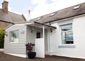 Small white and grey house with a sloping roof, front porch, and a potted plant near the entrance, set against a partly cloudy sky—an inviting Christian holiday cottage reminiscent of those found on Holy Island.
