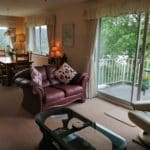 A cosy living room with a leather sofa, glass coffee table, and armchair awaits you in this Christian holiday flat. The inviting dining area features a wooden table and chairs, while sliding glass doors open to a balcony with outdoor seating overlooking the picturesque greenery of Bowness-on-Windermere.