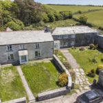 Aerial view of a stone farmhouse with green lawns, gardens, and a separate stone building, surrounded by trees and open fields under a blue sky. Ideal as a Christian retreat house in Cornwall, this peaceful setting also features a black car parked in the drive.