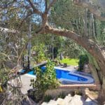 A small swimming pool and a hot tub are seen through the branches of a tree at this Christian holiday flat in Malaga, with stone steps in the foreground and green grass in the background on a sunny day.