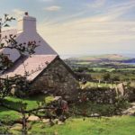 A charming white cottage, ideal as a Christian holiday cottage, nestles behind a stone wall on a grassy hillside overlooking the sea and a distant green peninsula in Pembrokeshire. Red berries grow on a branch in the foreground beneath a partly cloudy sky.