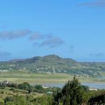 A scenic view of rolling green hills, scattered houses, and a river under a bright blue sky with a few clouds sets the perfect backdrop for a tranquil Christian holiday home in Donegal.