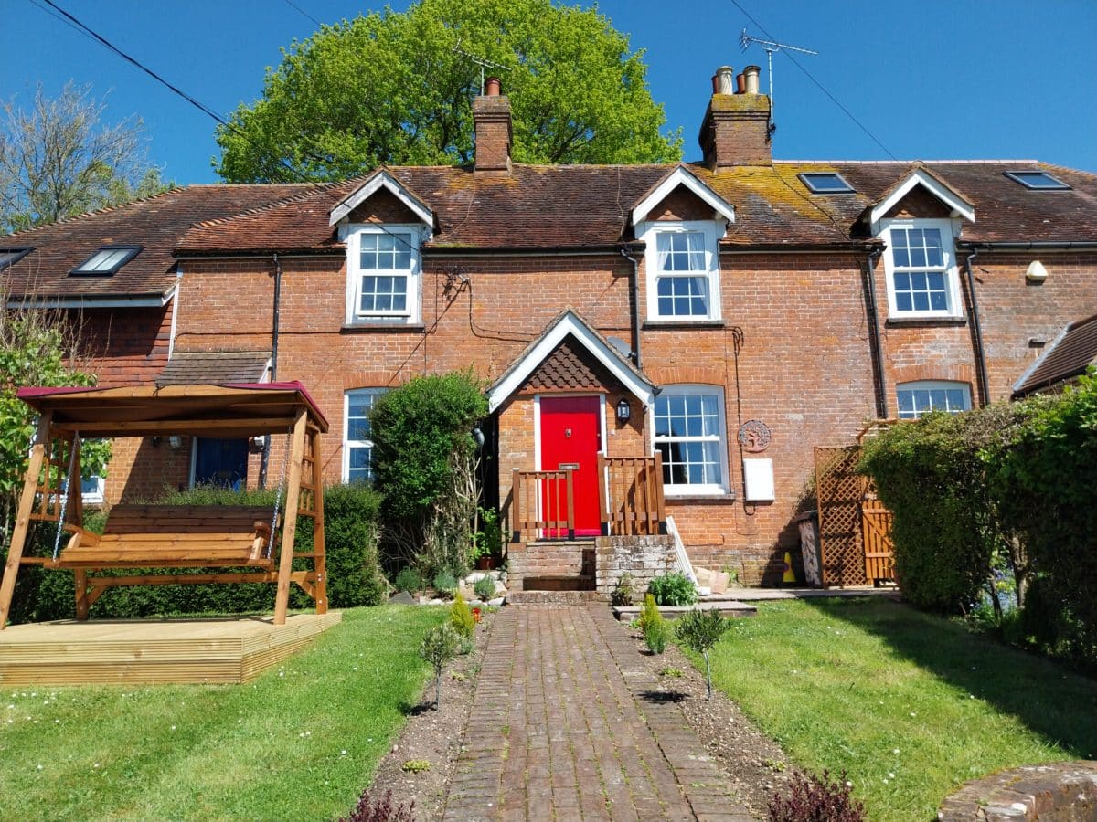 A charming brick house in Kent with a red front door, white window frames, and twin chimneys. Reminiscent of a classic Christian holiday cottage, it features a wooden swing on the left and a brick path winding through a lush green lawn to the entrance. Mature trees provide a picturesque backdrop.