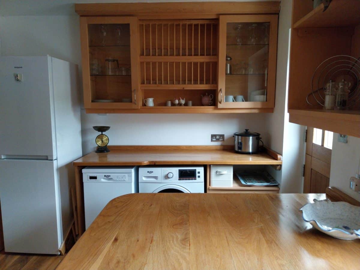 A kitchen in a charming Christian holiday cottage in Kent, featuring a wooden worktop, fridge, dishwasher, washing machine, slow cooker, scales, and wooden shelves with crockery and glasses.