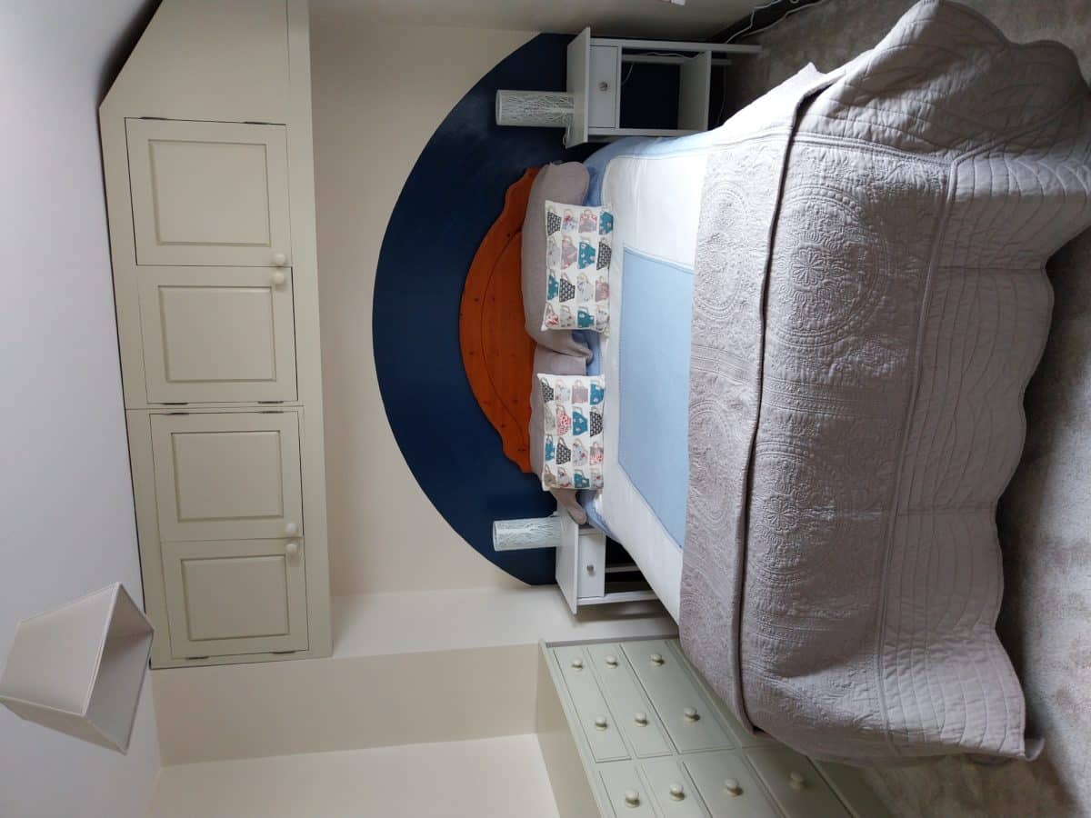 A neatly made bed with butterfly pillows, a blue and brown headboard, two bedside tables with lamps, overhead cupboards, and a chest of drawers in a cosy, softly lit bedroom evokes the inviting charm of a Christian holiday cottage in Kent.