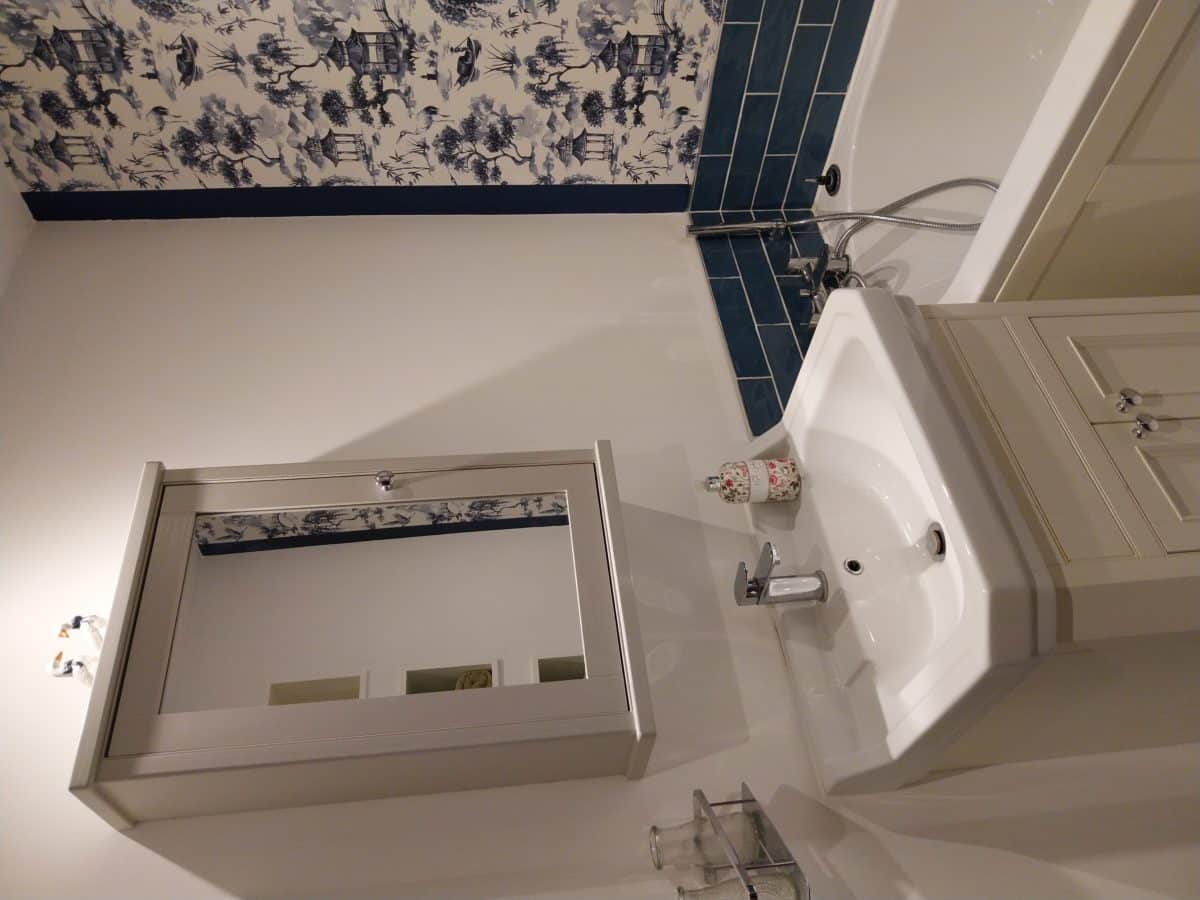 A bathroom in this charming Christian holiday cottage in Kent features a white basin with a cupboard, a mirrored medicine cabinet above, blue metro tiles, and a bath with blue-and-white Asian-inspired wallpaper on one wall.