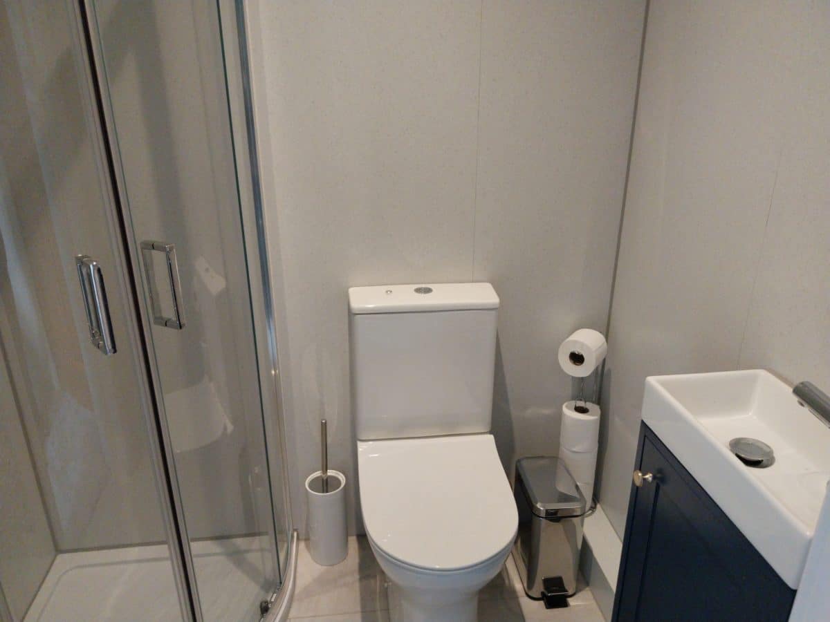 A small bathroom in this charming Christian holiday cottage in Kent features a shower, a toilet, a toilet brush, extra loo rolls, a rubbish bin, and a small washbasin with a cupboard underneath.