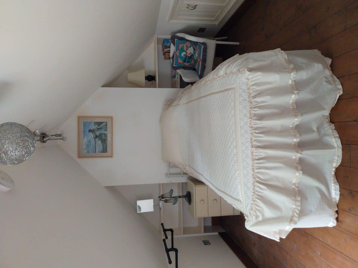 A cosy attic bedroom in a Christian holiday cottage in Kent, featuring a single bed in the centre with cream bedding, bedside table, lamp, armchair with a decorative cushion, wooden floor, and a painting on the wall above the bed.