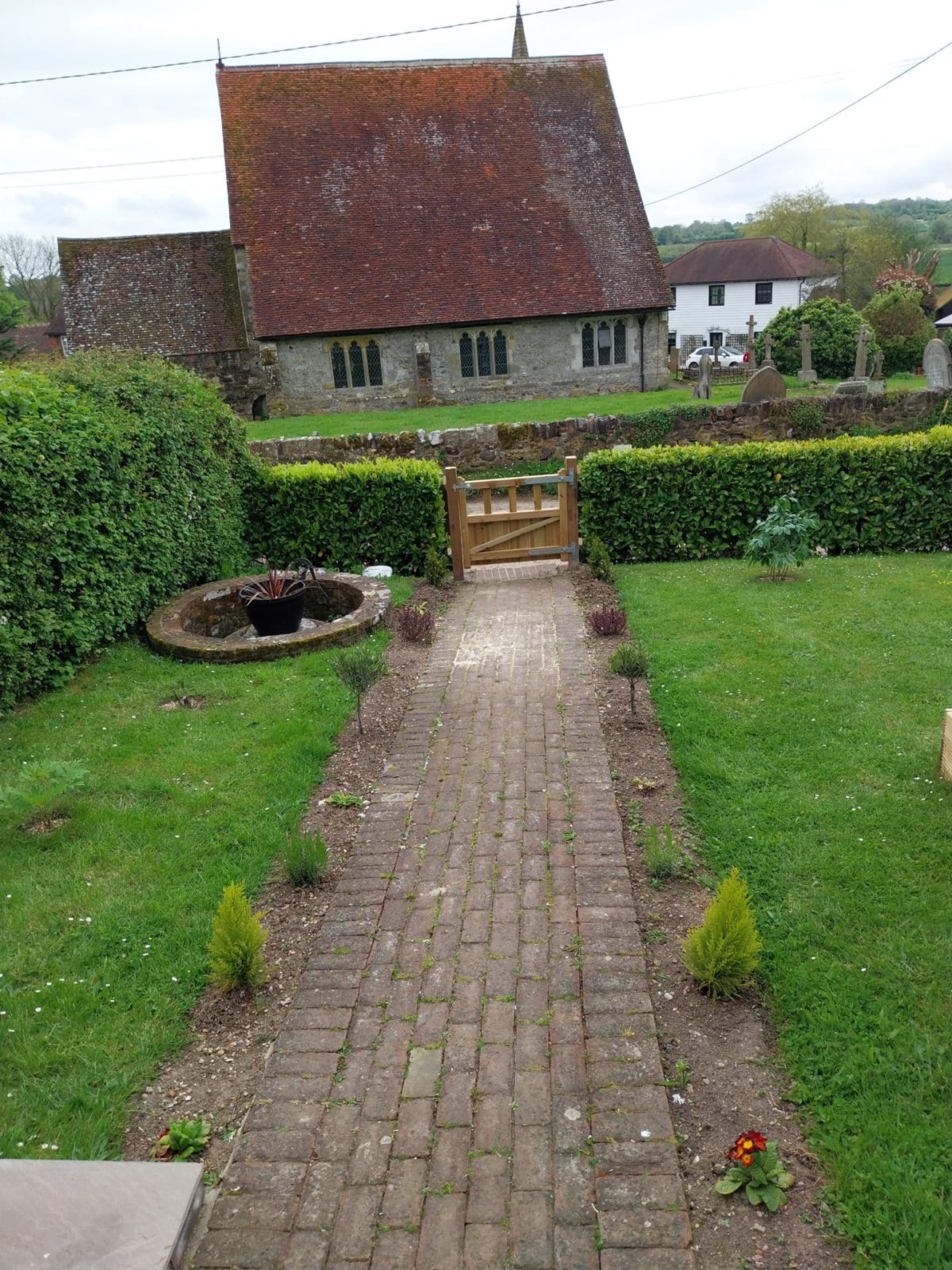 Brick path with small plants leads to a wooden gate, beyond which is an old stone church with a red-tiled roof. Set in the heart of Kent, the garden is bordered by green hedges and grass on both sides—an idyllic scene reminiscent of a classic Christian holiday cottage.