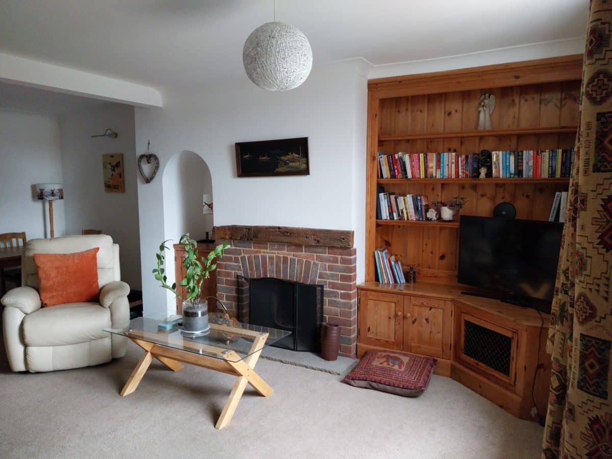 Cosy living room in a charming Christian holiday cottage in Kent, featuring a beige armchair, glass coffee table, brick fireplace, wooden bookcase with books and a TV, and a potted plant. Patterned curtains and wall art add a homely touch to this welcoming retreat.
