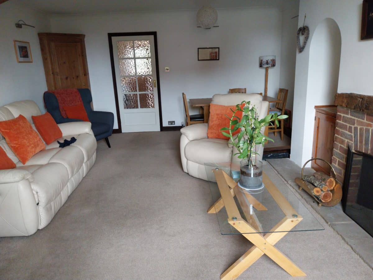 A cosy living room in a Christian holiday cottage in Kent, featuring beige sofas and orange cushions, a glass coffee table with a plant, a fireplace with firewood, and a dining table with chairs at the back. A closed wooden door and glass-paned door are visible.