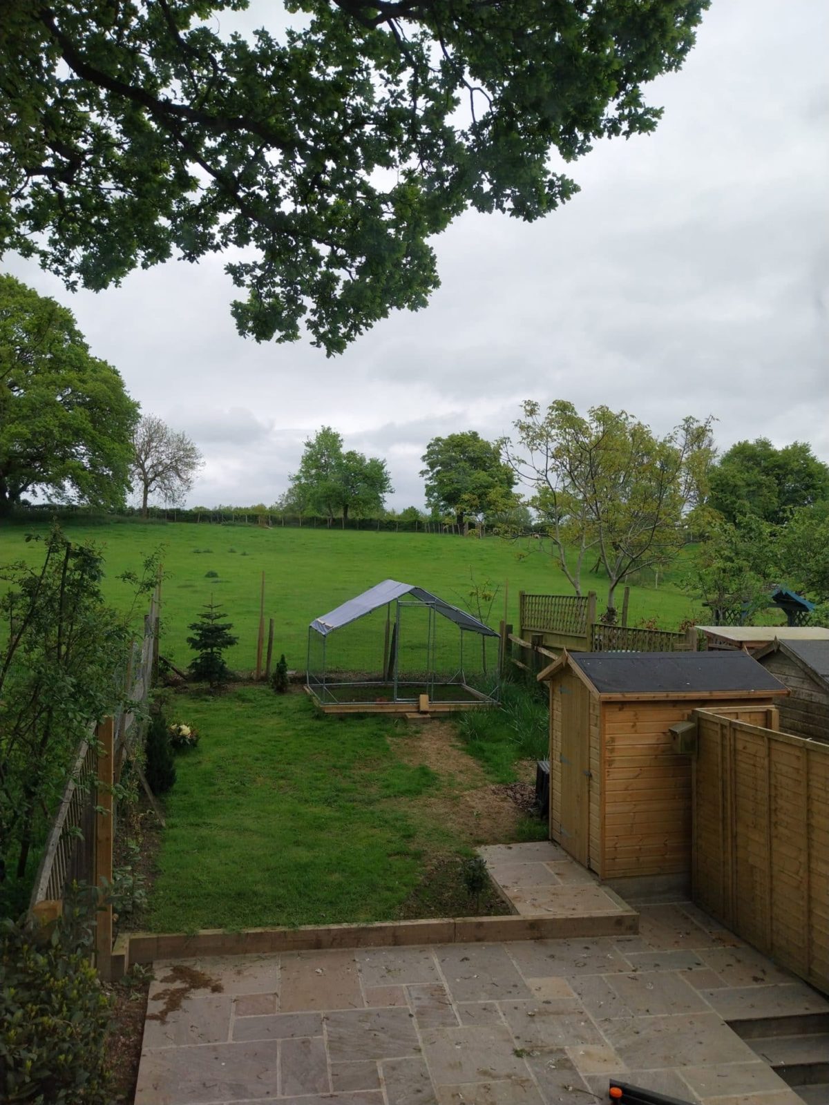 A back garden with a stone patio, wooden shed, fenced grassy area, and a metal-framed enclosure, reminiscent of a tranquil Christian holiday cottage in Kent, overlooking a green field with trees under a cloudy sky.