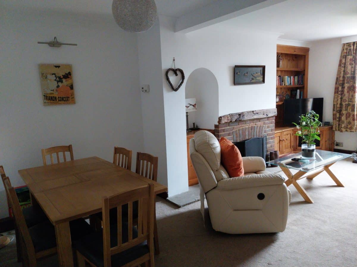 A cosy living-dining room in this Kent Christian holiday cottage features a wooden dining table and chairs, a white recliner, glass coffee table, TV, bookcase, wall art, and a brick fireplace. Light from a window brightens the room.
