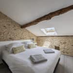 Cosy bedroom with stone walls, a large bed with white linen and yellow pillows, two bedside tables with lamps, folded towels, skylight, exposed wooden beam, and light wood flooring—perfect for a relaxing stay in a Dordogne Christian holiday gîte.