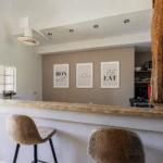 Modern kitchen with a wooden breakfast bar, two brown chairs, a rustic wooden pillar, and three framed art pieces on the wall that read BON, a heart symbol, and EAT. Neutral colours and minimalist decor are featured throughout this inviting Christian holiday gîte in the Dordogne.