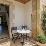 A small patio with a round table, two chairs, a wine bottle, and glasses sits outside a charming stone house with flowers nearby. Through the open door of this Dordogne Christian holiday gîte, a dining area and clock are visible inside.