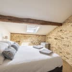 A cosy bedroom with a stone feature wall, wooden beam ceiling, skylight, neatly made bed with white linen and two patterned cushions, towels on the bed, and a small white cupboard—perfect for your stay at this charming Christian holiday gîte in Dordogne.
