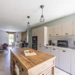 Modern kitchen with light cupboards, a wooden island, pendant lights, and small appliances on the worktop. In the background of this Dordogne Christian holiday gîte, a dining table and chairs sit near a window with a view of greenery.