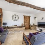 Cosy living room with stone walls, wooden beams, two grey sofas with purple cushions, a large wall clock, a TV on a white cabinet, and a rustic wooden door—perfect for relaxing in this charming Dordogne Christian holiday gîte.