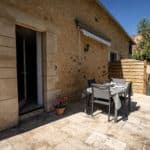 A stone patio with a dining table and four chairs, set for a meal, outside a rustic stone house in the heart of the Dordogne. Perfect for a Christian holiday gîte retreat, there are potted flowers by the door and a wooden privacy screen in the background, all under a clear blue sky.