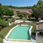Aerial view of a rectangular swimming pool surrounded by greenery, with a rustic stone house and shaded patio area in the background on a sunny day.