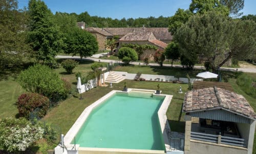 Aerial view of a rectangular swimming pool surrounded by greenery, with a rustic stone house and shaded patio area in the background on a sunny day.