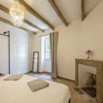 Bright bedroom with wooden ceiling beams, a bed with folded towels, a chandelier, a fireplace with a flower vase, a standing mirror, a curtain-covered window, and a clothes rail with hangers.