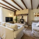Cosy living room with beige sofas, a wood-burning stove, exposed wooden ceiling beams, a TV, and large window. Walls are cream-coloured with light brown tiled floor and rustic decor accents.