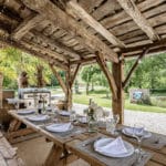 Rustic outdoor dining area with a wooden pergola, a set table for six, a brick barbecue, a nearby stainless steel barbecue, and lush greenery in the background.