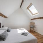 A cosy attic bedroom with exposed wooden beams, a skylight, a double bed with white linen and blue pillows, folded towels, and a wooden rocking chair on a hardwood floor.