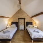 A cosy attic bedroom with two single beds, folded towels, a wooden floor, exposed beams, stone wall, chandelier, shelf, bedside tables, and lamps. Natural light enters from a skylight on the right.