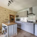 Modern kitchen with stone wall, small island, grey cabinets, gas cooker, mini-fridge, appliances, wall shelves, and tiled floor. Wine bottle and glasses sit on the island. Bright, clean, and organised space.