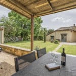 Covered patio with a table, wine bottle, glasses, book, and barbecue overlooks a grassy garden with trees and a rustic house in the background under a partly cloudy sky.