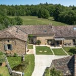 Aerial view of a rustic stone house with a tiled roof, green lawn, small pond, and surrounding trees under a blue sky.
