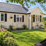 A single-storey yellow house with black shutters, white windows, a bicycle near the entrance, hanging flower baskets, and a lush green lawn bordered by colourful flowers under a sunny sky with some trees.