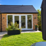 A modern brick home extension with large glass doors opens onto a sunny garden with green grass, a black planter with bushes, a potted plant, and part of a patio parasol and glass table visible.