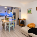 A cosy open-plan living room and kitchen with a beige sofa, colourful dining chairs around a wooden table, and soft lighting creating a warm atmosphere. Kitchen cupboards and LED lights are visible in the background.