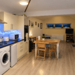 A cosy, modern kitchen and dining area with wooden floors, cream cupboards, a washing machine, fairy lights, a wooden dining table with chairs, and a living space with a TV and coat rack in the background.