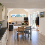 Bright open-plan kitchen and living room with light wood floors, cream cupboards, a dining table, a cosy sofa, TV, large windows, and glass doors opening to a garden.