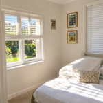 Sunlit bedroom with white walls, a neatly made bed, a small cushion, and a pink teddy bear. Framed pictures hang on the wall. Sunlight streams through two windows with white blinds, casting shadows on the bed.