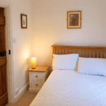 A cosy bedroom with a wooden bed, white bedding, two pillows, a bedside table with a lit lamp, and art on cream-coloured walls. A wooden door is on the left side of the room.