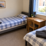 A tidy bedroom with two single beds in blue check bedding, each with a dark towel. There's a wooden desk and chair by the window, a potted plant, and a framed landscape picture on the wall.
