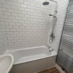A white-tiled bathroom with a bathtub, overhead shower, wall-mounted towel rail, and a washbasin. The floor is wood-patterned, and the walls feature glossy metro tiles.