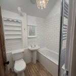 A small bathroom with wood-effect flooring, white metro tiles around a bath, a pedestal basin, a toilet, wall shelves, a window, and a shell chandelier hanging from the ceiling.