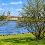 A stone building with a tower stands by a lake under a blue sky, seen through trees and green grass in the foreground.