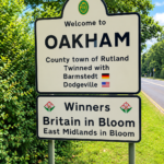 Road sign reads: Welcome to Oakham, County town of Rutland. Twinned with Barmstedt and Dodgeville. Winners Britain in Bloom, East Midlands in Bloom. Green trees and road in background.